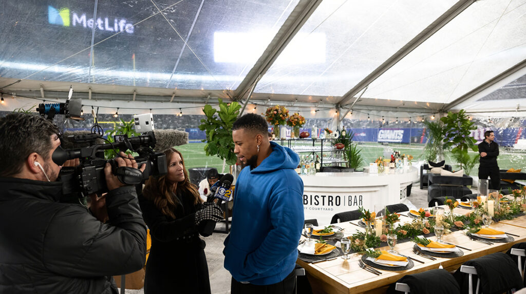 Saquon Barkley being interviewed by a reporter at the Courtyard by Marriott's Fansgiving event, with camera crews capturing the moment against the backdrop of the New York Giants stadium and a lavishly set dining area under a clear tent.