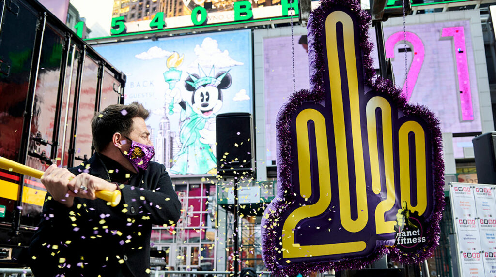 Amidst the towering skyscrapers of Times Square, a gigantic purple foam hand with "2020" on it. Pulling back on a yellow bat, preparing to swing. A cheerful Mickey Mouse graphic is visible on a nearby screen, along with a bright "21" display.